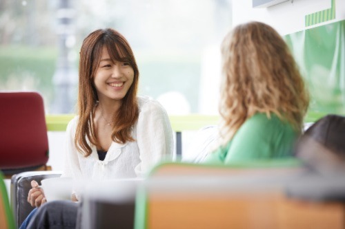 Two students smile and chat to each other in a University of Bristol building. One is wearing white and drinking from a coffee cup, the other is wearing green.